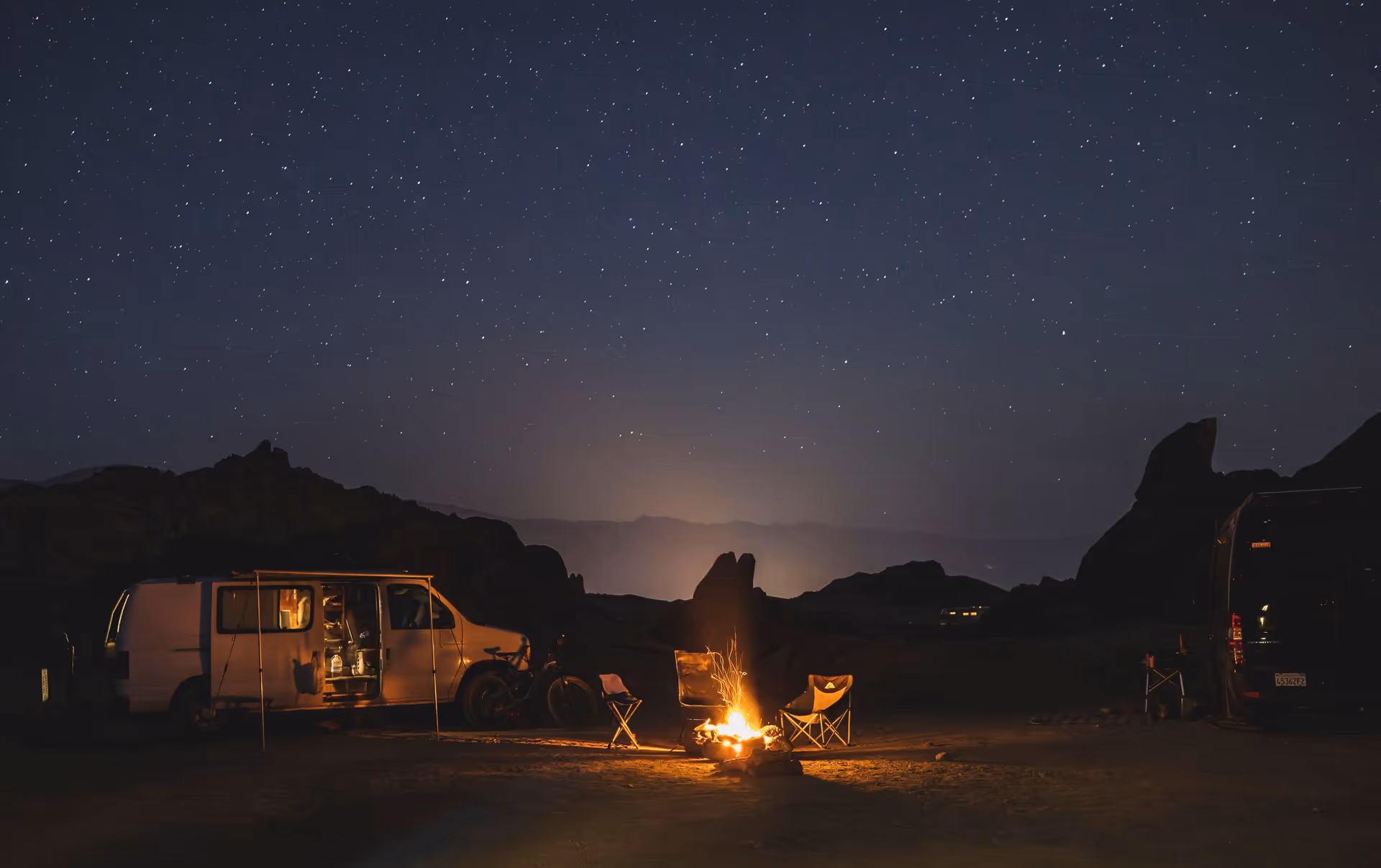 A camper van and a small campfire among boulders under a starry sky